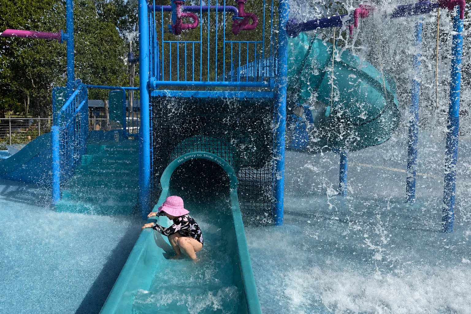 A child in a pink sunhat uses the small slide while water cascades over WetSide Water Park’s splash-play structure